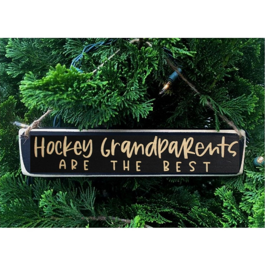 A rectangular black wooden hanging ornament with engraved lettering that reads 'Hockey Grandparents ARE THE BEST', displayed against green evergreen branches.