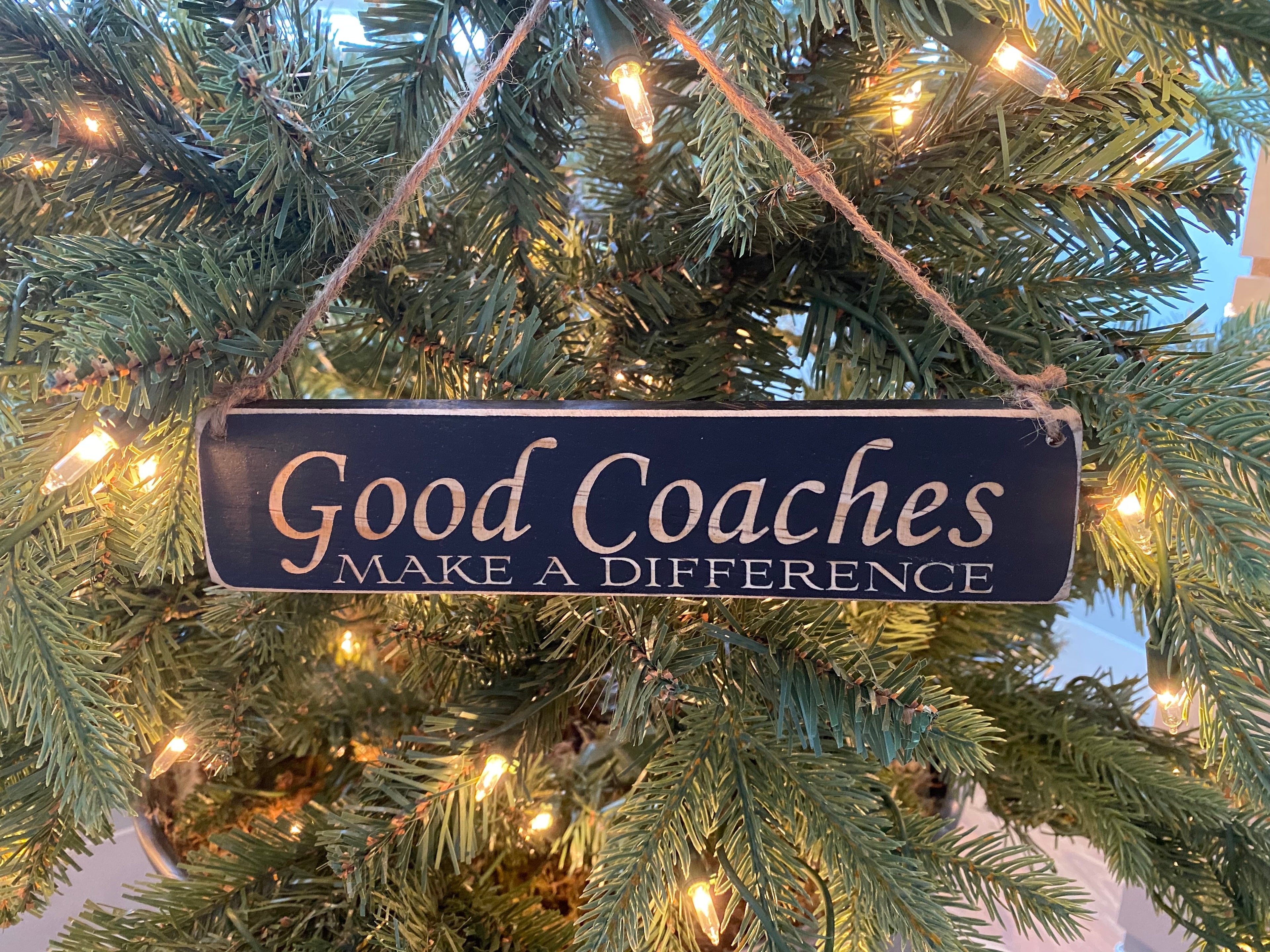 A rectangular dark blue wooden hanging ornament with the words 'Good Coaches MAKE A DIFFERENCE' in lasered lettering, suspended by a rope from a lit Christmas tree.