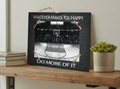 A rectangular black decorative sign with a rope handle, featuring the phrases 'WHATEVER MAKES YOU HAPPY' at the top and 'DO MORE OF IT' at the bottom, with a black-and-white basketball court photo in the center. The sign is displayed on a wooden shelf next to stacked books and a small potted succulent.