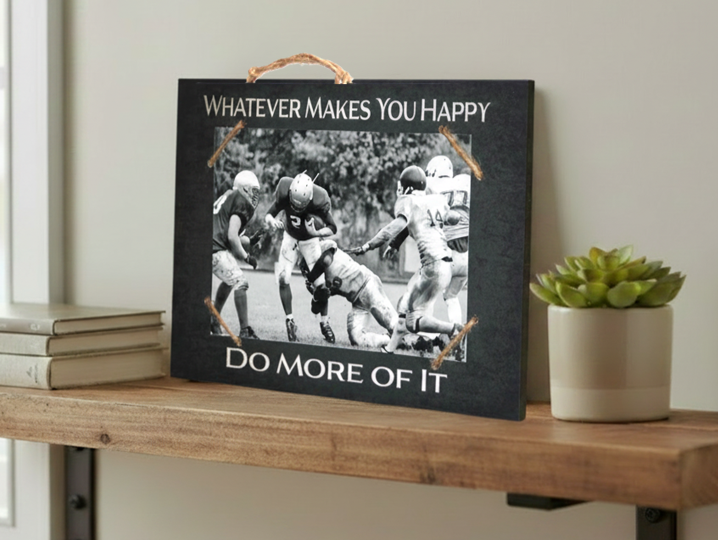 A rectangular black sign with a rustic wood-look finish sits on a wooden shelf. The sign has a rope hanger at the top and white text reading “WHATEVER MAKES YOU HAPPY” at the top and “DO MORE OF IT” at the bottom.