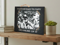 A rectangular black sign with a rustic wood-look finish sits on a wooden shelf. The sign has a rope hanger at the top and white text reading “WHATEVER MAKES YOU HAPPY” at the top and “DO MORE OF IT” at the bottom.