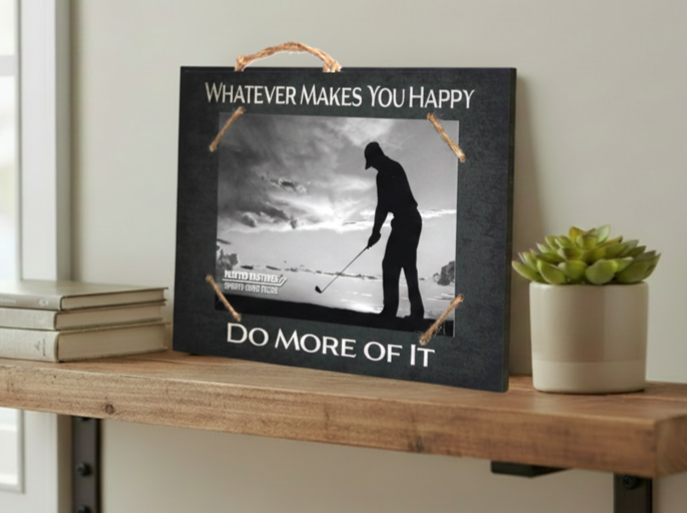 A rectangular black decorative sign with a rustic twine handle and corner ties, displayed on a wooden shelf. The sign features a black-and-white photo of a golfer and reads 'WHATEVER MAKES YOU HAPPY' at the top and 'DO MORE OF IT' at the bottom.