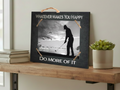 A rectangular black decorative sign with a rustic twine handle and corner ties, displayed on a wooden shelf. The sign features a black-and-white photo of a golfer and reads 'WHATEVER MAKES YOU HAPPY' at the top and 'DO MORE OF IT' at the bottom.