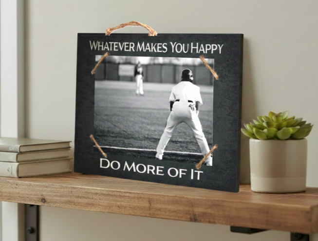 A rectangular black framed wall sign with a rope handle, featuring a black-and-white baseball player photo and the text 'WHATEVER MAKES YOU HAPPY' at the top and 'DO MORE OF IT' at the bottom. The sign is displayed on a wooden shelf next to books and a small potted succulent.