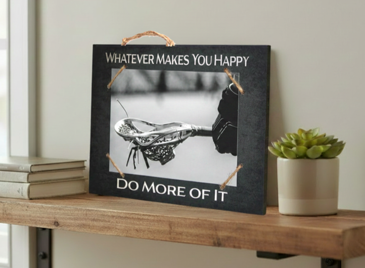 A rectangular black decorative sign with a rustic twine handle and corner ties, displayed on a wooden shelf. The sign features a black-and-white photo of a lacrosse head and reads 'WHATEVER MAKES YOU HAPPY' at the top and 'DO MORE OF IT' at the bottom.