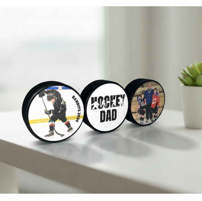 Three hockey pucks displayed on a white surface in a bright indoor setting. The center magnet reads 'HOCKEY DAD' in bold black text, while the side magnets show hockey players on ice.  The other two pucks show custom hockey photos.