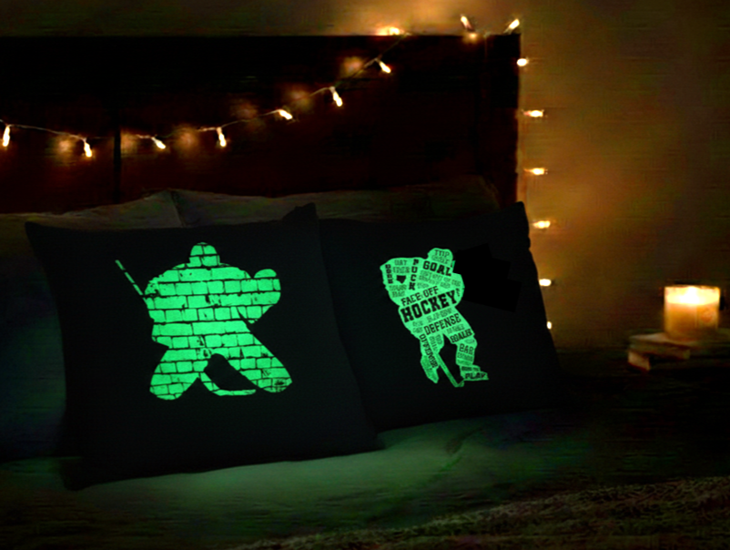 Two glow-in-the-dark pillows on a bed in a dimly lit bedroom. One pillow shows a hockey goalie silhouette made of brick textures, and the other shows a hockey player silhouette made of words like 'GOAL', 'SKATE', and 'HOCKEY'.
