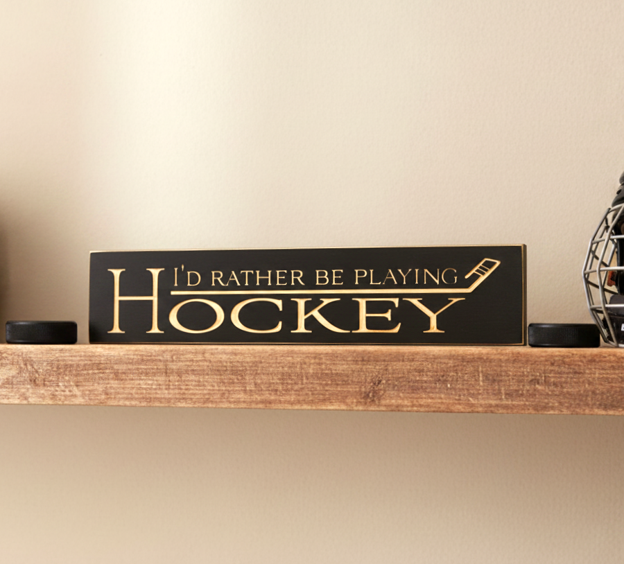 A rectangular black wooden sign with engraved lettering that reads 'I'D RATHER BE PLAYING HOCKEY' sits on a rustic wooden shelf. The sign features a hockey stick graphic and is flanked by two black hockey pucks, with a hockey mask partially visible on the right.