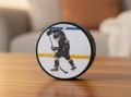A custom hockey photo puck with a custom printed graphic of a hockey player in a black jersey, standing on a wooden table with a blurred living room background.