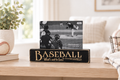 A black wooden baseball-themed sign on a light wood table in a bright living room setting. The sign reads 'BASEBALL What's not to love?' in engraved lettering, with a photo of a baseball game displayed on top.