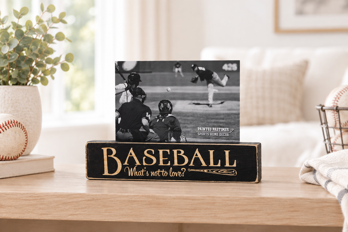 A black wooden baseball-themed sign on a light wood table in a bright living room setting. The sign reads 'BASEBALL What's not to love?' in engraved lettering, with a photo of a baseball game displayed on top.