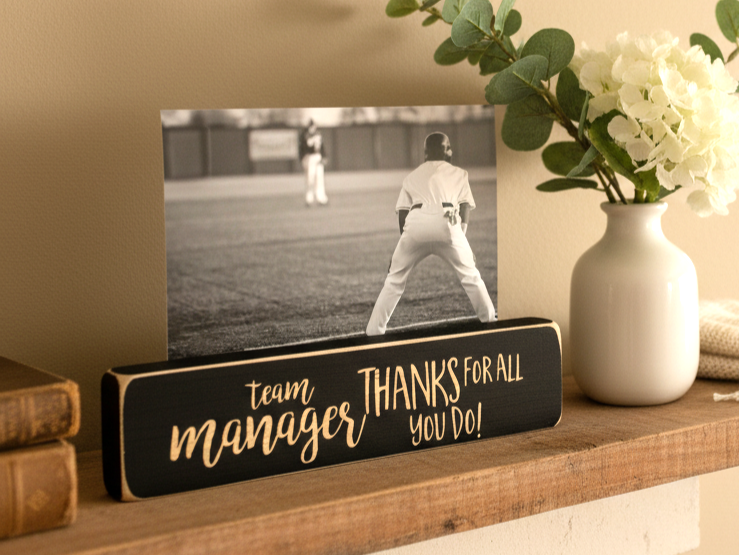 A black wooden photo block sign with lasered lettering that reads 'team manager THANKS FOR ALL YOU DO!' displayed on a wooden shelf next to a white vase with flowers and a framed baseball photo.