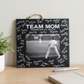 A personalized black chalkboard-style wall plaque hanging on a light wood table, featuring a central black-and-white baseball photo of a batter and white text reading 'TEAM MOM Thanks for all you do' with signatures around the border. A small plant, marker, and notebook are visible nearby.