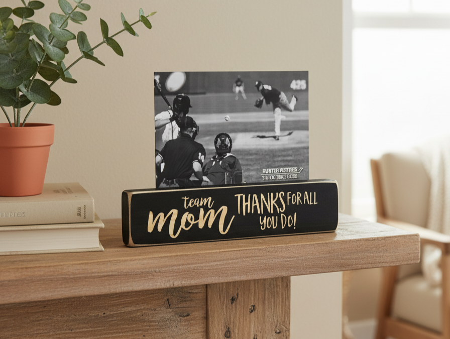 A black rectangular wooden sign with lasered lettering that reads 'team mom THANKS FOR ALL YOU DO!' sits on a wooden table in a home setting, holding a black-and-white baseball photo print. A potted plant and books are visible on the left, and a chair is softly blurred in the background.
