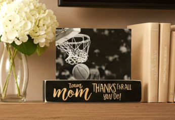 A rectangular black plaque or sign on a wooden shelf, featuring a black-and-white basketball court scene with a basketball near the hoop and lasered text reading 'Thanks for all you do!'. The sign is styled on a shelf beside white flowers in a glass vase and beige books.