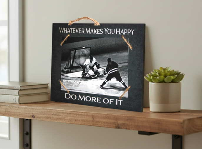 A rectangular black decorative sign with a rustic twine handle and corner ties, displayed on a wooden shelf. The sign features a black-and-white photo of a hockey and reads 'WHATEVER MAKES YOU HAPPY' at the top and 'DO MORE OF IT' at the bottom.