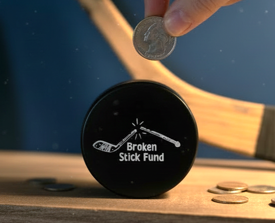 A black circular puck-shaped coin holder with a white hockey stick graphic and the text 'Broken Stick Fund' sits on a wooden surface. A hand is placing a coin into the holder, with additional coins scattered around and a hockey stick blurred in the background.