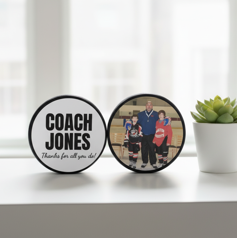 Two round custom hockey pucks displayed on a white shelf. One puck is white with black text reading 'COACH JONES' and 'Thanks for all you do!', the other is a custom photo.