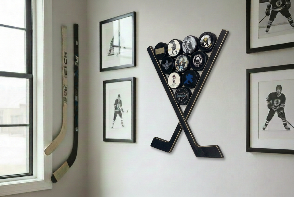 Two black wooden hockey sticks crossed to form a "V" area which displays hockey pucks.  This hangs on a wall amongst frames hockey photos and hockey sticks in a room setting with light pouring through a window.