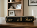 A rectangular black wooden sign with engraved lettering that reads 'Crawl... Walk... Skate!