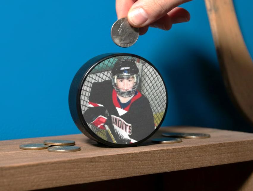 A custom hockey puck bank sits on a wooden shelf, featuring a printed custom photo of a hockey player. A hand is shown placing a coin into the slot on top of the puck, with other coins resting on the shelf below.