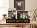 A decorative black wooden sign with carved lettering reading 'DREAMS HAPPEN HERE' sits on a wooden table in a softly lit living room. Two black-and-white football-themed photo prints are displayed on top of the sign.
