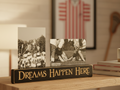 A rectangular black wooden sign with carved lettering that reads 'DREAMS HAPPEN HERE' sits on a wooden shelf in a warm indoor setting. Two small black-and-white photos are propped behind it on a shelf.