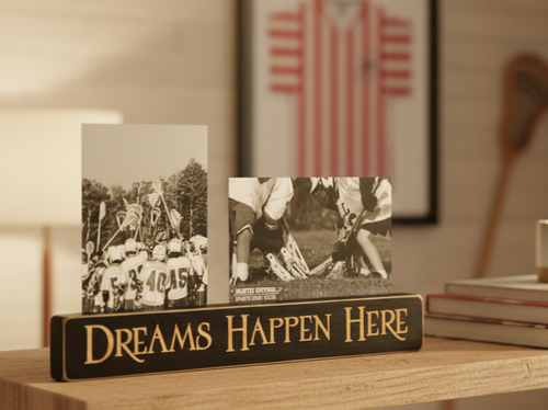A rectangular black wooden sign with carved lettering that reads 'DREAMS HAPPEN HERE' sits on a wooden shelf in a warm indoor setting. Two small black-and-white photos are propped behind it on a shelf.