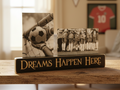 A wooden tabletop display with a black plaque reading “DREAMS HAPPEN HERE” in carved lettering, holding two vertical black-and-white photos: one of a goalkeeper catching a soccer ball and one of a group of soccer players huddled together. The scene is set in a warm indoor room with blurred background elements, including a framed red jersey with the number 10.