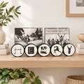 A set of 5 connected hockey pucks, spelling out the custom name "HENRY" with unique hockey letters.   This displays 2 hockey photos and is sitting on a natural wood table with plants and books in a cozy room with light pouring in.