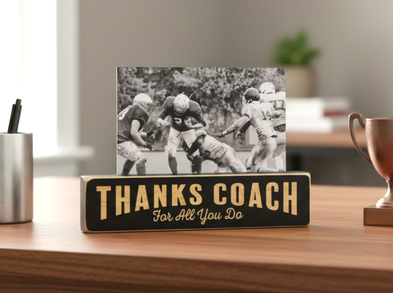 A rectangular black wooden sign on a wooden desk with the words “THANKS COACH For All You Do” in lasered lettering, holding a small black-and-white football photo behind it. The scene is styled in an office or home setting with a pen cup, blurred books, and a trophy in the background.