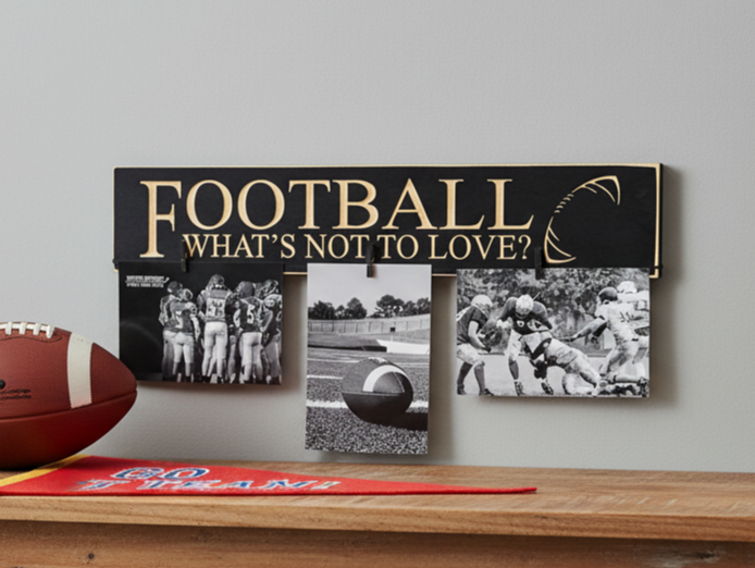 A wooden football-themed photo display sign with the text 'FOOTBALL WHAT'S NOT TO LOVE?' in carved lettering on a black background, shown mounted on a wall above a wooden surface. Three black-and-white football photos are clipped to the sign, and a brown football and a red-and-gold banner are partially visible in the foreground.