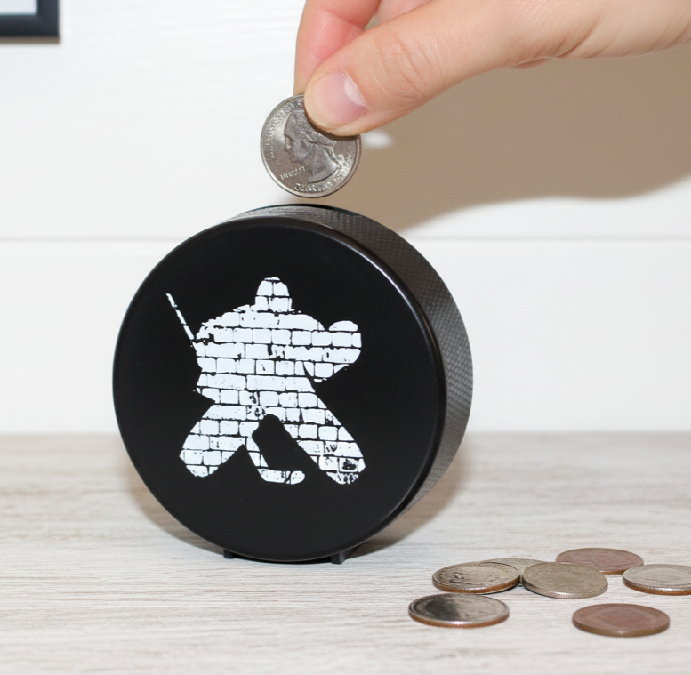 A black round piggy bank shaped like a hockey puck with a white hockey goalie made of bricks graphic, shown on a light wood surface with a hand inserting a coin.
