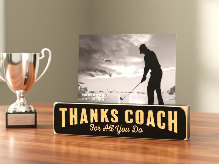 A rectangular black wooden sign with lasered lettering that reads 'THANKS COACH For All You Do' sits on a wooden table next to a silver trophy cup. A black-and-white golf photo card is propped behind the sign.