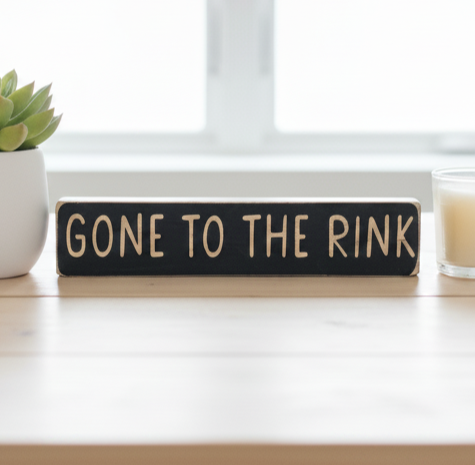 A rectangular black wooden sign with the phrase 'GONE TO THE RINK' in engraved lettering, displayed on a light wood tabletop with a small potted succulent on the left and a candle on the right.
