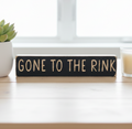 A rectangular black wooden sign with the phrase 'GONE TO THE RINK' in engraved lettering, displayed on a light wood tabletop with a small potted succulent on the left and a candle on the right.