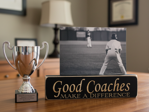 A black wooden sign with lasered lettering that reads 'Good Coaches MAKE A DIFFERENCE' sits on a wooden table next to a silver trophy cup. Behind the sign is a black-and-white photo frame showing a baseball player on a field.