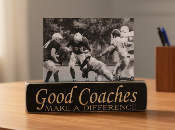 A rectangular black plaque on a wooden desk with the words 'Good Coaches MAKE A DIFFERENCE' in lasered lettering, holding a black-and-white football photo print. A blurred indoor setting with a pen holder is visible in the background.