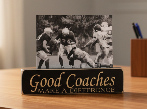 A rectangular black plaque on a wooden desk with the words 'Good Coaches MAKE A DIFFERENCE' in lasered lettering, holding a black-and-white football photo print. A blurred indoor setting with a pen holder is visible in the background.