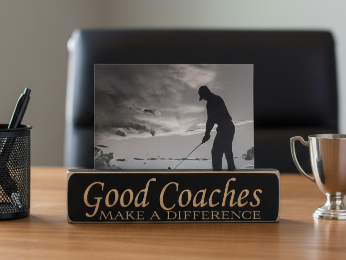 A rectangular wooden sign with the phrase 'Good Coaches MAKE A DIFFERENCE' in lasered lettering sits on a wooden desk. Behind it is a black desk chair, and the sign holds a black-and-white photo of a golfer.