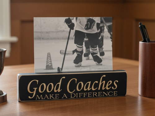 A rectangular black wooden sign with lasered lettering that reads 'Good Coaches MAKE A DIFFERENCE' sits on a wooden table, holding a black-and-white hockey photo behind it. A pen cup and other desk items are visible in the background.
