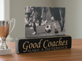 A rectangular black wooden plaque with lasered lettering that reads 'Good Coaches MAKE A DIFFERENCE' sits on a wooden table next to a silver trophy cup. Behind the plaque is a black-and-white photo of soccer players running toward a ball.