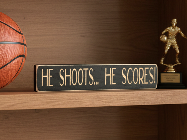 A wooden sign with engraving "He Shoots... He Scores" in a basketball setting shelf