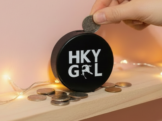 A hockey puck bank with  HKY GRL in  white lettering sits on a wooden shelf, with a hand inserting a coin into the top slot. Several coins are scattered around the box, and warm fairy lights glow softly in the background against a soft pink wall.