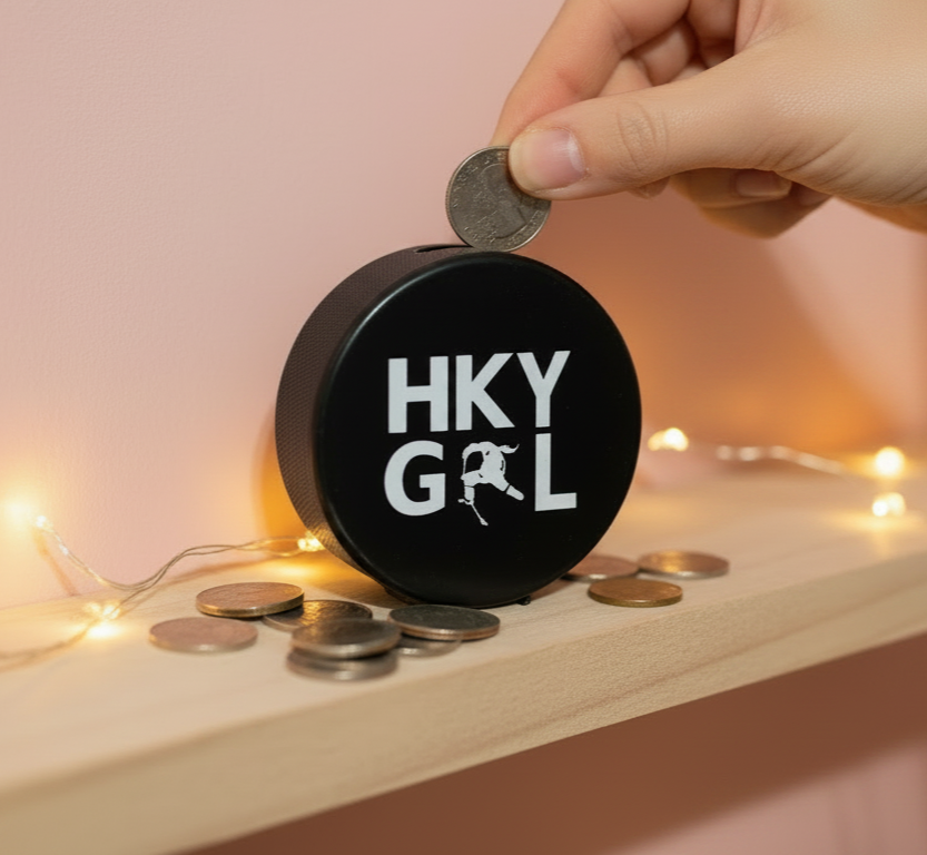 A hockey puck bank 'HKY GRL' printed in white on the front, shown on a wooden shelf with coins and a hand inserting a coin. Warm fairy lights glow in the background against a soft pink wall.