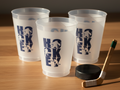 Three frosted plastic cups with a blue 'HOCKEY' graphic printed on them, arranged on a wooden tabletop alongside a black hockey puck and a wooden hockey stick.