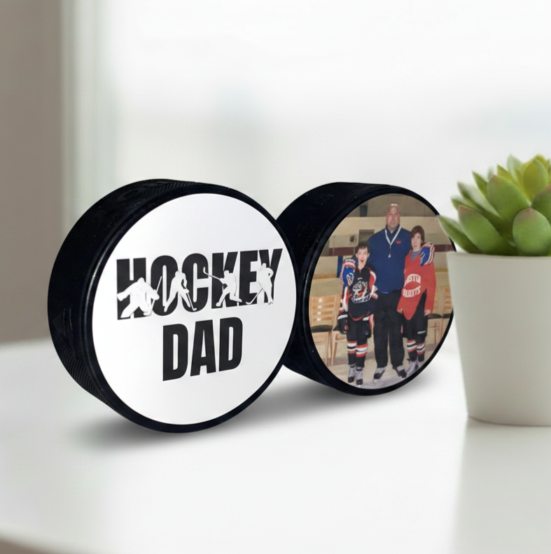 Two hockey pucks displayed on a white tabletop. One puck shows the text 'HOCKEY DAD' with hockey player silhouettes, and the other shows a printed family photo of a father and children on an ice rink.