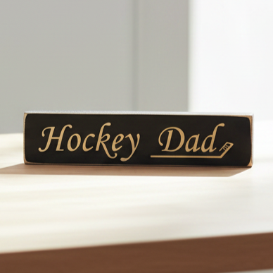 A rectangular black wooden sign with engraved lettering that reads 'Hockey Dad' resting on a light wood surface against a soft gray background.