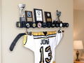 A black wooden hockey trophy shelf mounted on a beige wall, holding several framed awards and plaques and trophies.  The edges are holding special hockey pucks and a hockey jersey hanging below from a hockey stick.