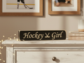 A rectangular black wooden sign with 'Hockey Girl' in engraved lettering sits on a distressed white dresser in a cozy room setting with framed pictures on the wall and warm fairy lights.